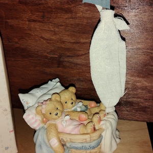 May include: A figurine of two tan teddy bears resting in a woven basket boat with a white fabric sail and a blue triangular flag. The basket features a banner with the text Smooth Sailing, and the bears are nestled against a white pillow.