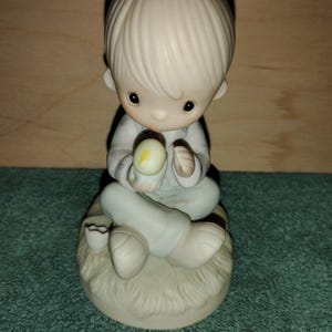 May include: A porcelain figurine of a child sitting cross-legged on a textured base. The child wears light-colored pajamas with a small blue floral pattern and holds a small yellow and white egg, with a broken eggshell fragment nearby.