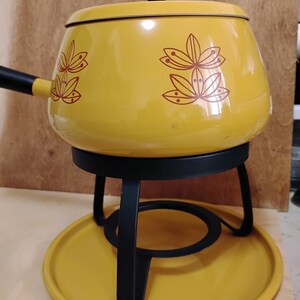 May include: A yellow enamel fondue pot with a matching lid and a black handle. The pot features a repeating dark red floral pattern. It rests on a black metal stand with a circular base, which is set upon a matching yellow circular metal tray.