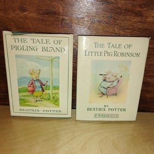 May include: Two hardcover books by Beatrix Potter titled The Tale of Pigling Bland and The Tale of Little Pig Robinson. Each book features a cream-colored dust jacket with a central color illustration of a pig character and green text.