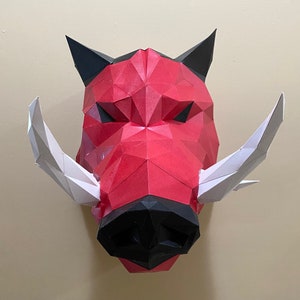 May include: A red and black papercraft boar head with white tusks. The boar's face is made of folded paper and has a fierce expression.