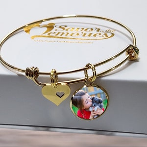 May include: A gold-toned expandable wire bangle bracelet featuring two charms: a solid gold heart with a small cutout heart and a circular pendant displaying a colour photograph of a child holding a small puppy. The bracelet is metallic gold.