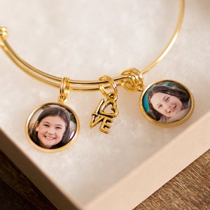 May include: A gold-toned expandable bangle bracelet featuring two circular photo charms displaying portraits of a smiling child and a central gold-toned charm with the word LOVE and a heart shape, all presented in a square gift box.