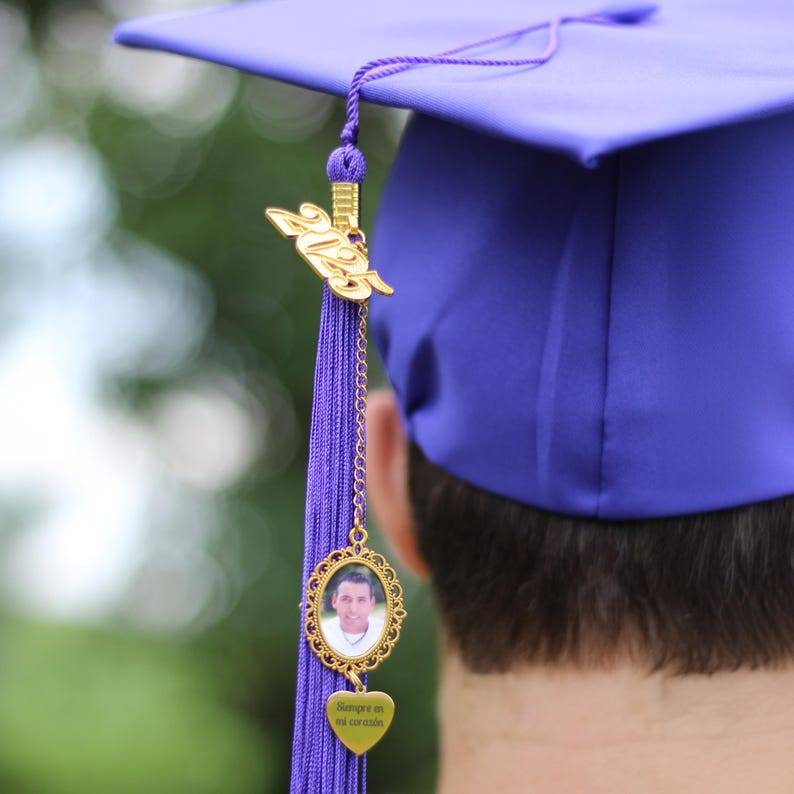 Puede incluir: Un gorro de graduaci&oacute;n morado con una borla dorada que presenta un dije de foto y un dije de coraz&oacute;n con el texto "Siempre en mi coraz&oacute;n".