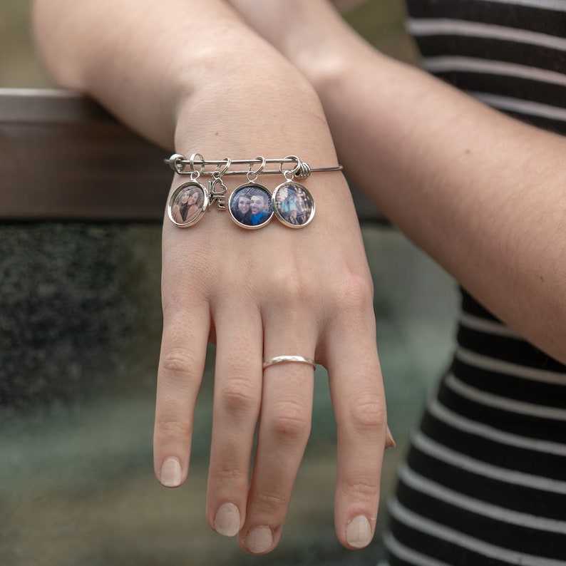 May include: A silver-toned expandable wire bangle bracelet featuring three circular photo charms and one small silver-toned heart-shaped charm. The bracelet is worn on a wrist, and a simple, plain silver-toned band ring is worn on the ring finger.