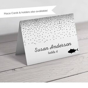 Op de afbeelding: Witte plaatskaart met een zilveren glitterhartpatroon. De tekst "Susan Anderson tafel 4" is op de kaart gedrukt. Een zwart silhouet van een vis bevindt zich in de rechterbenedenhoek.
