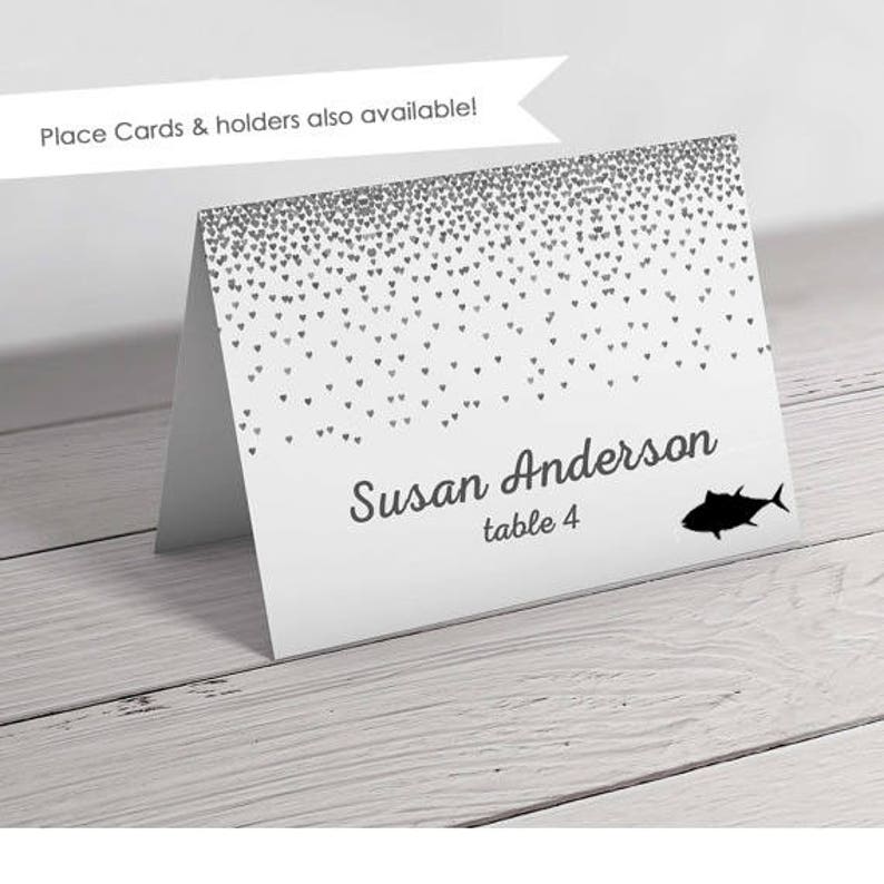 Op de afbeelding: Witte plaatskaart met een zilveren glitterhartpatroon. De tekst "Susan Anderson tafel 4" is op de kaart gedrukt. Een zwart silhouet van een vis bevindt zich in de rechterbenedenhoek.