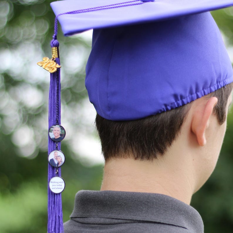 Puede incluir: Un gorro de graduaci&oacute;n morado con una borla que presenta un amuleto dorado 2023 y tres amuletos de fotos redondos. La borla cuelga del gorro y las fotos son visibles.