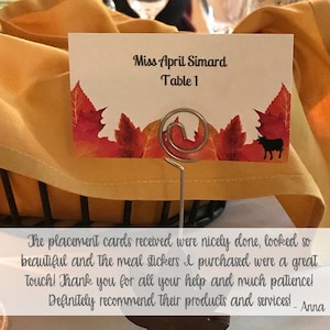 Op de afbeelding: Een witte kaart met de tekst "Miss April Simard Table 1" erop gedrukt. De kaart is versierd met een herfstthema met rode en oranje bladeren. Een metalen standaard houdt de kaart rechtop.