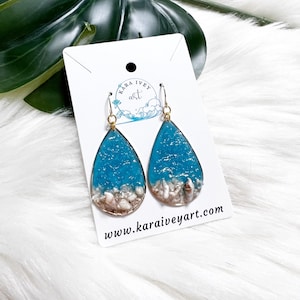 May include: Teal blue resin earrings with white and gold seashells embedded in the resin. The earrings are teardrop shaped and have gold hooks. The earrings are on a white card with the text "Kara Ivey Art" and "www.karaiveyart.com".