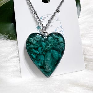 May include: A silver chain necklace with a heart-shaped pendant filled with teal green resin and small white and green pieces. The pendant is about 2 inches in diameter.