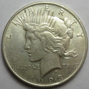 May include: A silver 1923 Peace dollar coin. The obverse features a profile of Lady Liberty with rays emanating from her crown, the word "LIBERTY" above, and "IN GOD WE TRUST" below. The year "1923" is at the bottom.