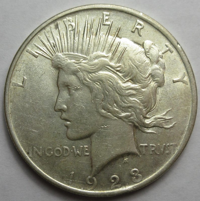 May include: A silver 1923 Peace dollar coin. The obverse features a profile of Lady Liberty with rays emanating from her crown, the word "LIBERTY" above, and "IN GOD WE TRUST" below. The year "1923" is at the bottom.