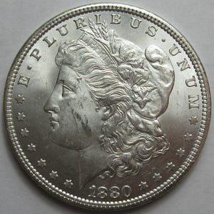 May include: A silver coin with a detailed profile of a woman wearing a cap, surrounded by the inscription "E PLURIBUS UNUM" and stars. The year "1880" is visible at the bottom. The coin has a detailed, textured surface.