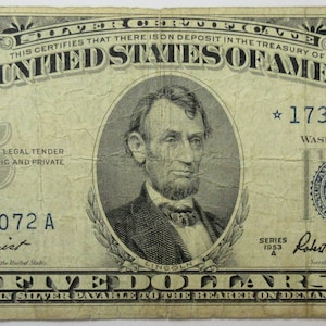 May include: A vintage five dollar bill featuring a portrait of Abraham Lincoln. The bill is dated 1923 and has the serial number *17355072 A. The text "THE UNITED STATES OF AMERICA" is printed in bold at the top of the bill.