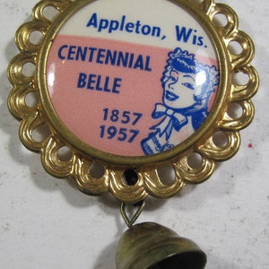 Może przedstawiać: Zabytkowa, złota metalowa przypinka z falistą krawędzią, z napisem "Appleton, Wis. CENTENNIAL BELLE 1857 1957" i niebiesko-białą ilustracją. Mały dzwonek wisi poniżej.