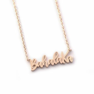 Pode incluir: Um delicado colar dourado com a palavra "Bahalaka" em escrita cursiva. O colar apresenta uma corrente fina e um pingente com o nome. A joia &eacute; exibida sobre um fundo branco.