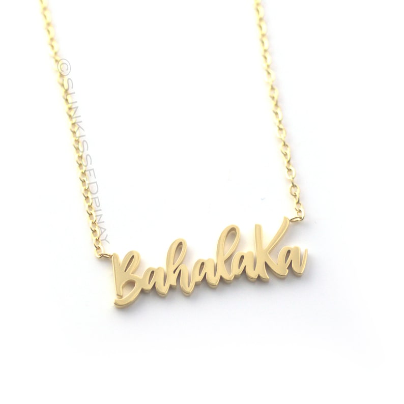 Pode incluir: Um colar dourado com a palavra "Bahalaka" em fonte cursiva. A corrente &eacute; delicada e o pingente est&aacute; centralizado. O fundo &eacute; branco.