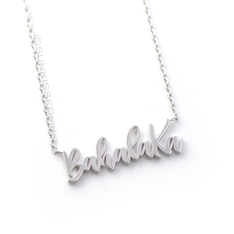 Pode incluir: Um colar prateado com a palavra "Bahalaka" em escrita cursiva. O colar est&aacute; pendurado em uma corrente fina contra um fundo branco. O nome da marca "SunKissedPinay" est&aacute; impresso no lado esquerdo da imagem.