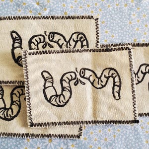 May include: A collection of beige fabric patches with black worm illustrations. Each patch has a black stitched border. The patches are arranged on a light blue floral patterned background.