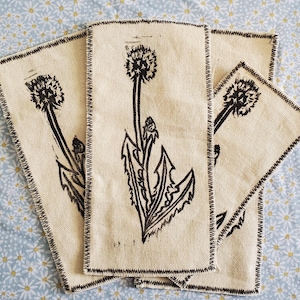 May include: Three rectangular, off-white fabric napkins with a black dandelion print. The napkins have a black stitched border and are arranged on a light blue floral patterned surface. The dandelion design includes the flower head, stem, and leaves.
