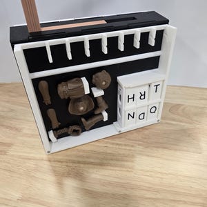 Op de afbeelding: Een witte en zwarte rechthoekige doos met diverse bruine 3D-geprinte objecten, waaronder een pistool, laarzen en een hoofd. De doos bevat ook witte lettertegels met de tekst "HRT NQ". Een houten potlood is bovenop geplaatst.