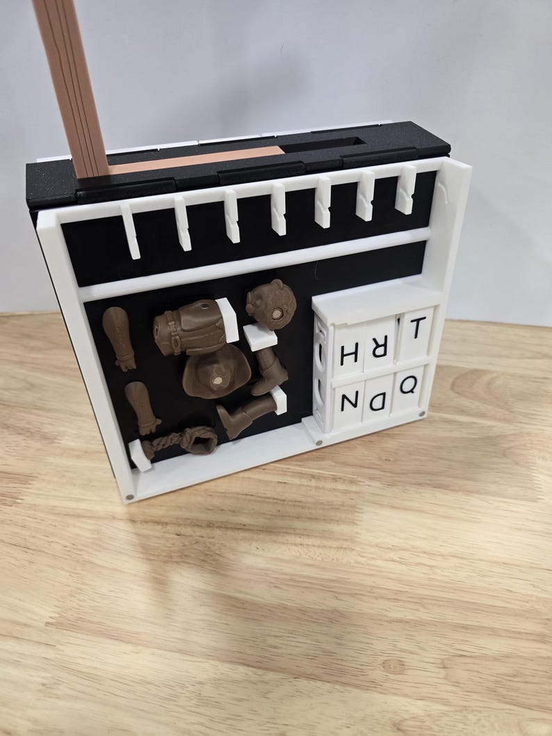 Op de afbeelding: Een witte en zwarte rechthoekige doos met diverse bruine 3D-geprinte objecten, waaronder een pistool, laarzen en een hoofd. De doos bevat ook witte lettertegels met de tekst "HRT NQ". Een houten potlood is bovenop geplaatst.