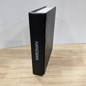 Op de afbeelding: Een zwart boek met het woord "HANGMAN" verticaal in het wit gedrukt op de rug. Het boek heeft een witte bovenrand en staat rechtop op een houten oppervlak.