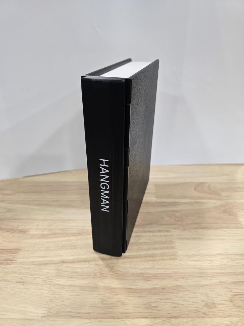 Op de afbeelding: Een zwart boek met het woord "HANGMAN" verticaal in het wit gedrukt op de rug. Het boek heeft een witte bovenrand en staat rechtop op een houten oppervlak.