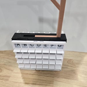 Op de afbeelding: Een witte, gelaagde, alfabetische display met individuele opklapbare compartimenten. De bovenkant heeft een zwart-wit gedeelte met een houten constructie die lijkt op een galg. De letters A tot en met G zijn zichtbaar.