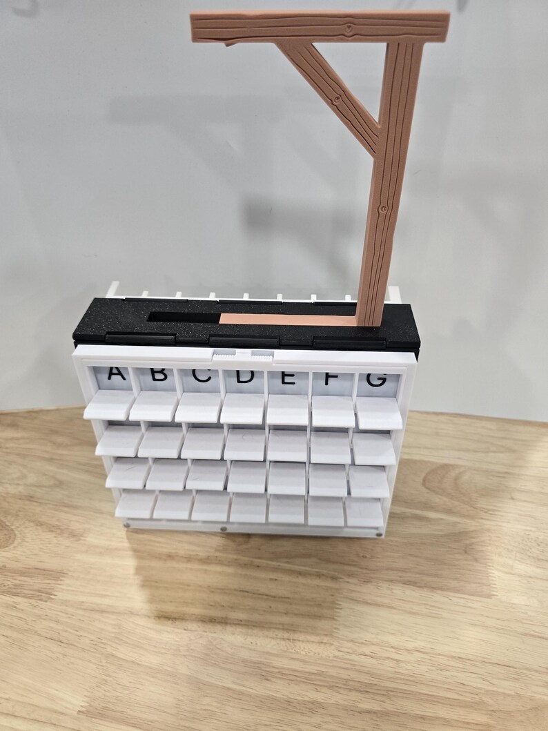 Op de afbeelding: Een witte, gelaagde, alfabetische display met individuele opklapbare compartimenten. De bovenkant heeft een zwart-wit gedeelte met een houten constructie die lijkt op een galg. De letters A tot en met G zijn zichtbaar.