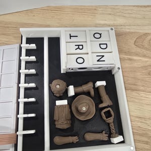 Op de afbeelding: Een witte en zwarte puzzel met de letters "T R O" en "O D N" zichtbaar. De puzzel bevat bruine miniatuurvoorwerpen, waaronder een hoed, jas en laarzen, gerangschikt op een zwart oppervlak in een wit frame. De puzzel staat op een houten oppervlak.