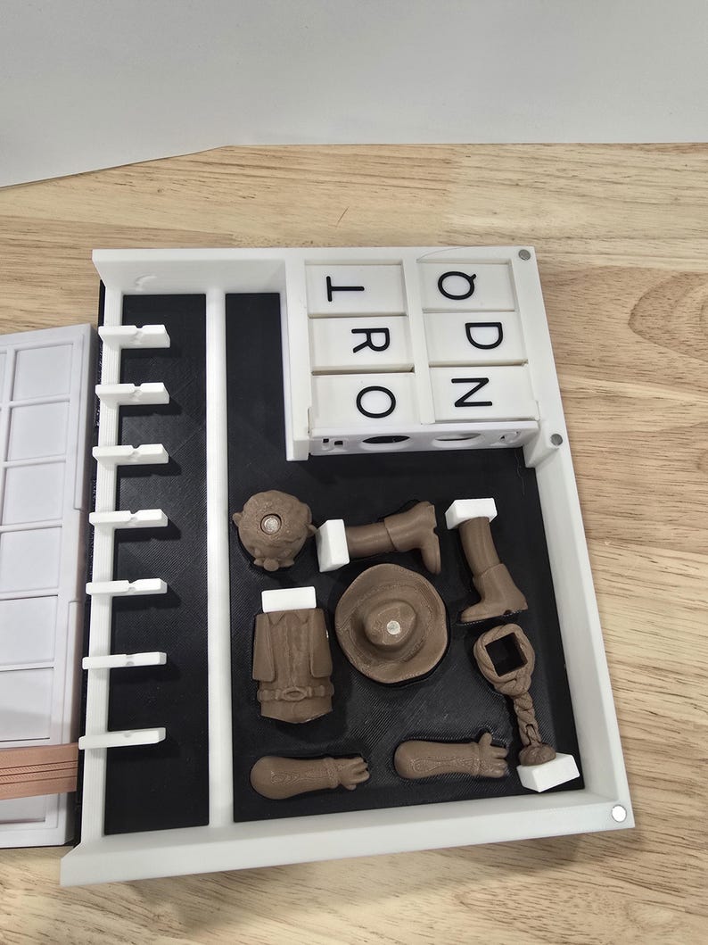 Op de afbeelding: Een witte en zwarte puzzel met de letters "T R O" en "O D N" zichtbaar. De puzzel bevat bruine miniatuurvoorwerpen, waaronder een hoed, jas en laarzen, gerangschikt op een zwart oppervlak in een wit frame. De puzzel staat op een houten oppervlak.