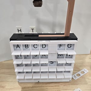 Op de afbeelding: Een wit woordspel met meerdere niveaus en tegels met letters. Het spel bevat een bruine houten steun en een klein bruin hoedvormig stuk dat aan een touw hangt. Extra lettertegels liggen in de buurt verspreid.