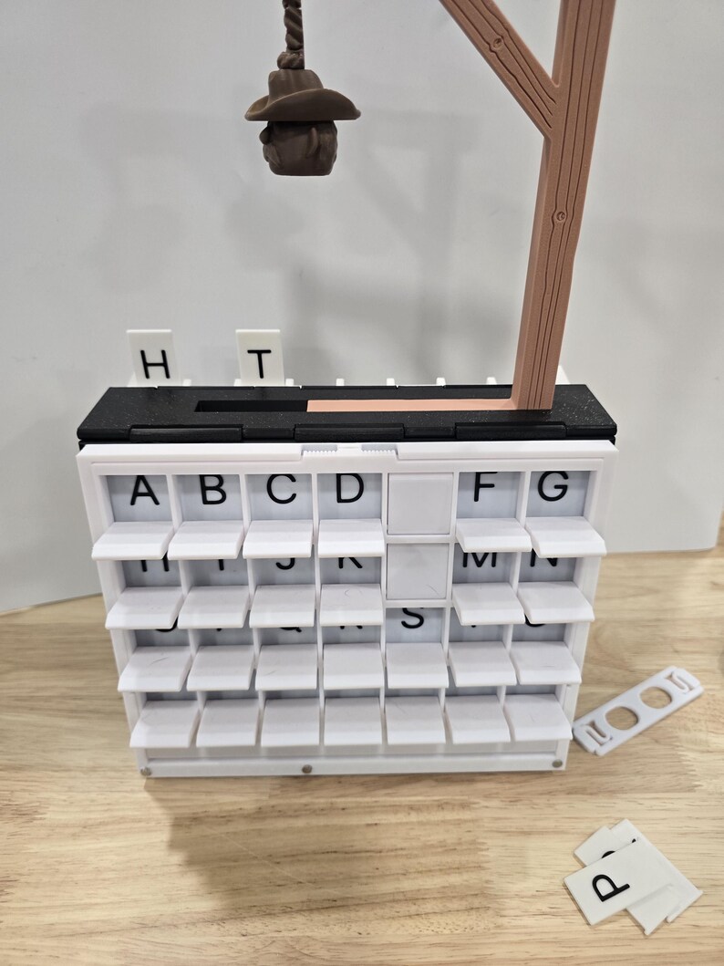 Op de afbeelding: Een wit woordspel met meerdere niveaus en tegels met letters. Het spel bevat een bruine houten steun en een klein bruin hoedvormig stuk dat aan een touw hangt. Extra lettertegels liggen in de buurt verspreid.