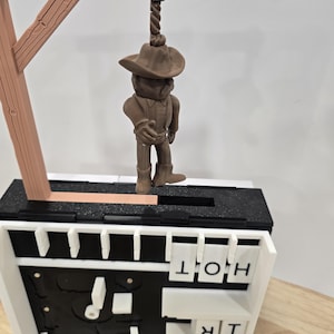 Op de afbeelding: Een bruin cowboyfiguur hangt aan een galg. Het figuur draagt een hoed, een overhemd en laarzen. De galg is van bruin hout. De basis is zwart en wit met de letters "HOT" en "TRQ".