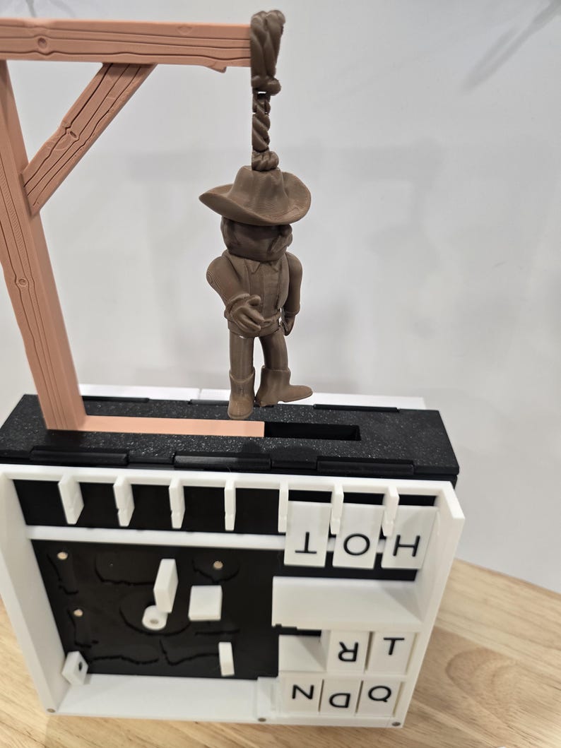 Op de afbeelding: Een bruin cowboyfiguur hangt aan een galg. Het figuur draagt een hoed, een overhemd en laarzen. De galg is van bruin hout. De basis is zwart en wit met de letters "HOT" en "TRQ".