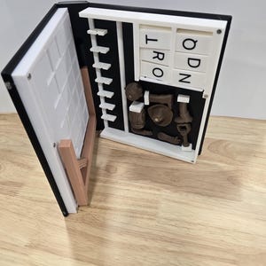 Op de afbeelding: Een open 3D-geprint boek met een zwarte rug en witte pagina's. De linkerpagina heeft een wit deurontwerp en de rechterpagina een raster met de letters "T", "R", "O", "Q", "D" en "N". Verschillende bruine miniatuur objecten worden getoond.