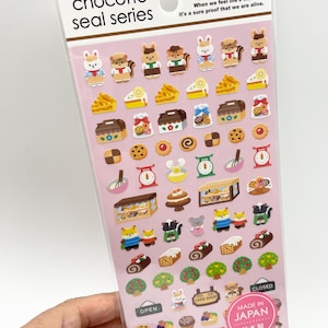 以下が含まれることがあります： Chocotto seal seriesのステッカーシート。淡いピンクの背景に、動物、ケーキ、クッキー、キッチンツール、小さな建物のイラストが描かれています。上部には「When we feel life's warmth, it's a sure proof that we are alive.」というテキストが記載されています。