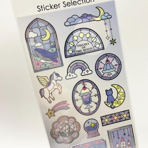 以下が含まれることがあります： Sticker SelectionとMade in Japanと書かれたステッカーシート。窓の中のクジラ、月の満ち欠け、翼のあるユニコーン、三日月に乗った猫、城、白鳥、その他様々なパステルカラーの天体モチーフのイラストが描かれています。