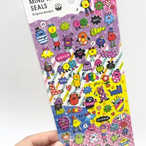 May include: A sheet of Mind Wave Seals Puchi Monster stickers featuring a variety of colourful, small cartoon characters in diverse shapes and expressions. The stickers are arranged on a background with purple, yellow, and diagonal striped sections.