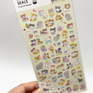 Peut inclure: Une planche d'autocollants Mind Wave Seals présentant de nombreuses petites illustrations colorées de chatons dans diverses poses, portant des accessoires comme des nœuds, des couronnes et des chapeaux, parsemées de cœurs, d'étoiles et de cadeaux.