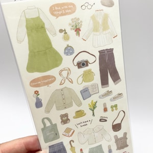 Peut inclure: Une planche d'autocollants intitulée Sticker Selection présentant des illustrations de vêtements, d'accessoires et d'objets, dont une robe verte, un pantalon marron, un appareil photo, un ours en peluche, des fleurs et des bulles de texte.