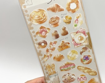 May include: A clear plastic sheet of adhesive stickers featuring watercolor illustrations of various baked goods, including bread, croissants, pastries, and sandwiches. The header text reads Sketch sticker, with additional handwritten notes bread, good day, and yummy.