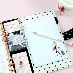 May include: A white planner with black polka dots and a gold ring binder. The planner has a black and white sticker sheet with a ballerina design, a pink and white sticker sheet with a cupcake design, and a white notepad with the words "Chic Notes" printed in pink. A white pencil with a gold tip is lying on the notepad. The planner also has a black and white polka dot strap with the words "Unleash Your Creativity" printed in gold.