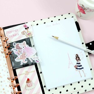 May include: A white planner with black polka dots and a rose gold coil binding. The planner has a sticker sheet with a pink and white design and a note pad with the words "Chic Notes" written on it. A white pencil with a gold eraser is lying on the note pad.