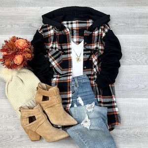 May include: A black and white plaid flannel shirt with a black hooded sweatshirt attached to the sleeves. The shirt is worn over a white ribbed tank top and blue jeans. The outfit is accessorized with a gold necklace and brown suede ankle boots.