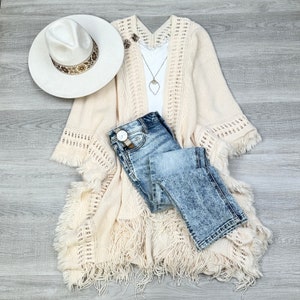 May include: A cream-colored crochet cardigan with fringe detailing, a pair of light blue denim jeans, and a white hat with a brown band.