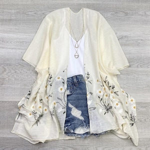 May include: A white sheer kimono with embroidered daisies and a delicate floral pattern. The kimono has short sleeves and a relaxed fit. It is paired with denim shorts.