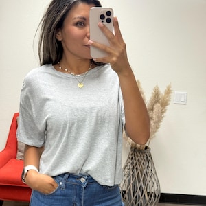 May include: A light gray t-shirt with short sleeves. The shirt is paired with blue jeans. A gold heart pendant necklace and a white watch are visible. The person is holding a phone.